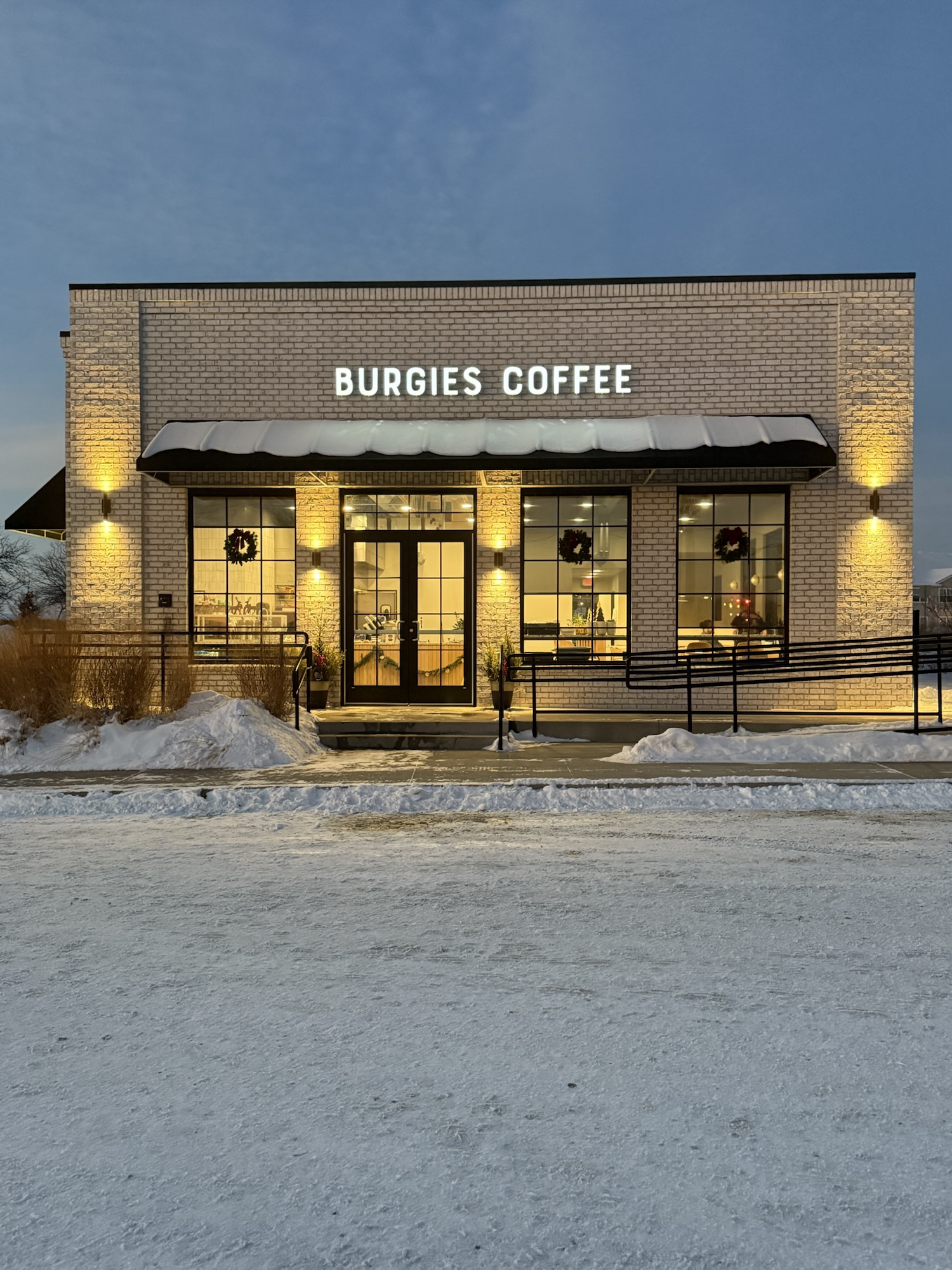 Exterior of Burgies Coffee North Ames on Stange Road on a snowy winter evening with warm lights glowing through the windows